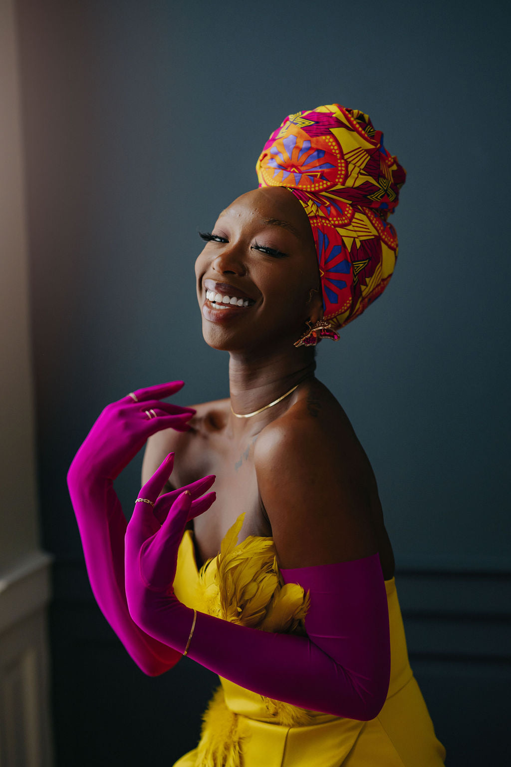 Woman wearing a colorful headwrap and bright yellow dress with pink gloves against a dark background